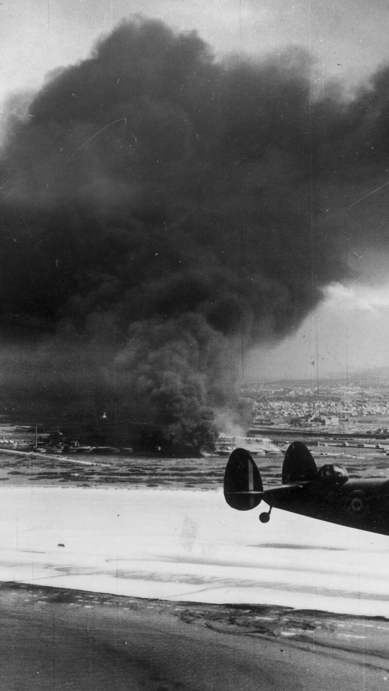 A Coastal Command Lockhead Hudson flying past burning oil tanks on Dunkirk beach
