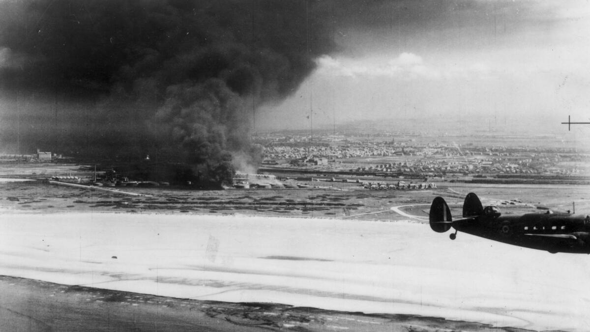 A Coastal Command Lockhead Hudson flying past burning oil tanks on Dunkirk beach